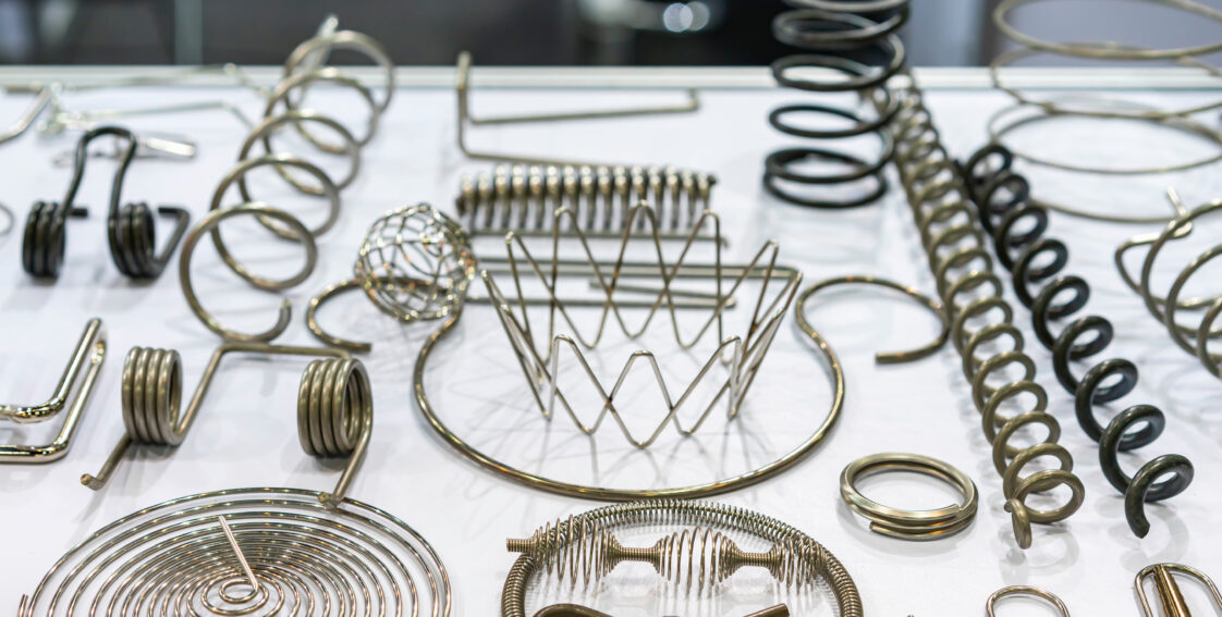 Multiple wire forms are laid out on a white tabletop. The wire forms are made from different grades of steel and feature varying designs, including coils